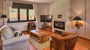 Un salón con sofá, sillón, mesa de centro, televisor, chimenea y una mesita con lámpara y flores.