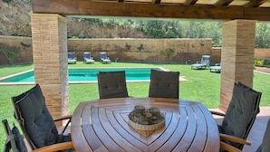 Una mesa de madera con sillas bajo una pérgola, con vista a una piscina y sillones de jardín.