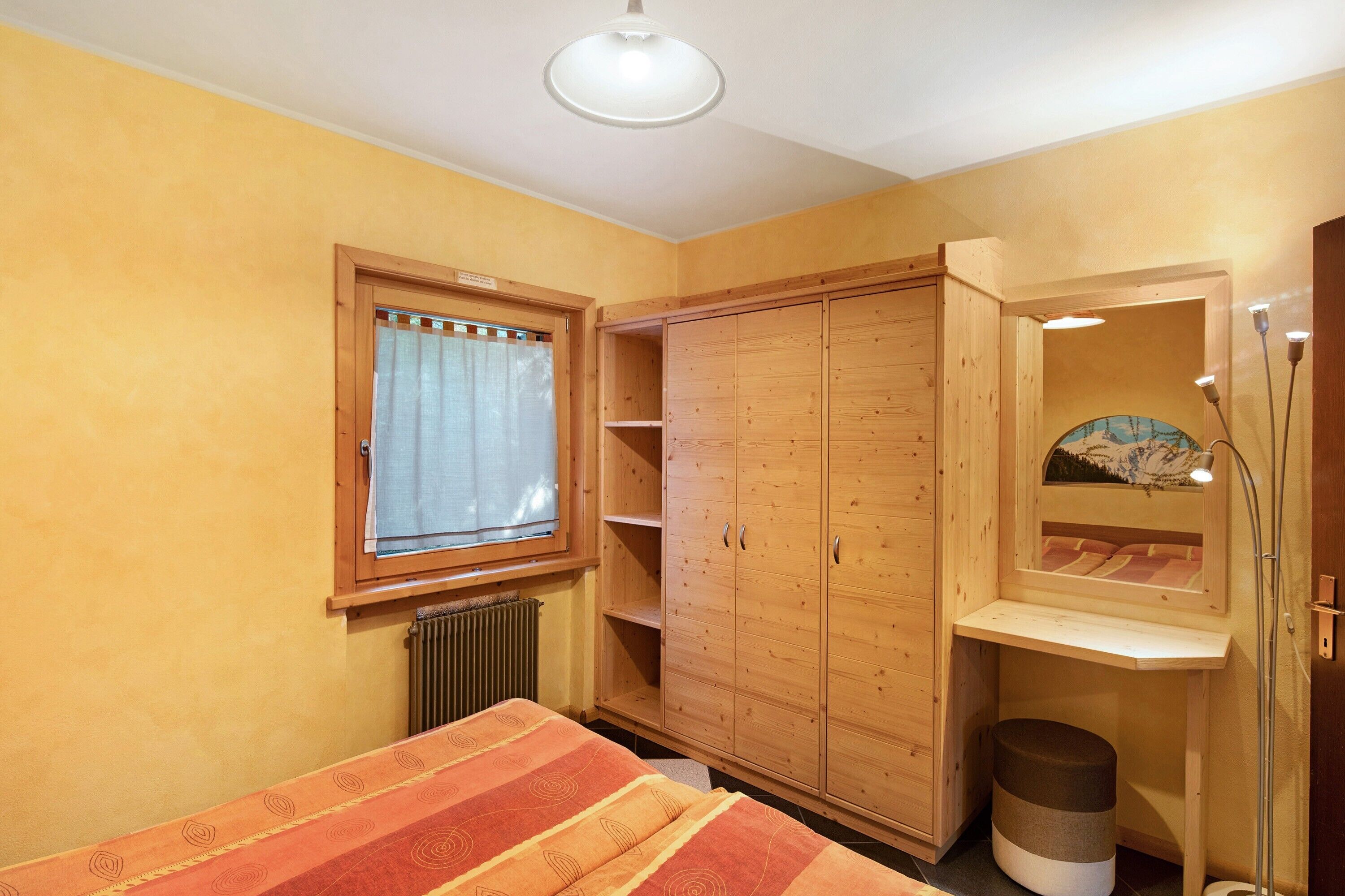 A bedroom with a bed, wardrobe, window, radiator, and a small stool.
