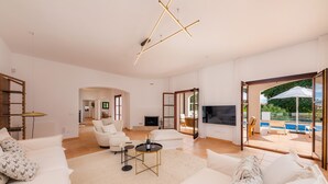 A spacious living room with a large sofa, a coffee table, and a TV. There is a sliding glass door leading to an outdoor area with a pool. - Villa 'Font de Sa Cala' with Sea View, Wi-Fi and Air Conditioning (Provensals)