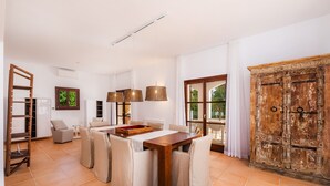A spacious living area with a wooden dining table, cushioned chairs, a sofa, a wooden cabinet, and a large window with curtains. - Villa 'Font de Sa Cala' with Sea View, Wi-Fi and Air Conditioning (Provensals)