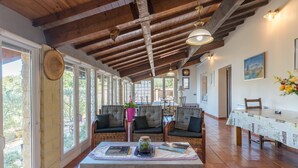 A spacious room with a high wooden ceiling, large windows, wicker furniture, and a dining table set with books and a jar.