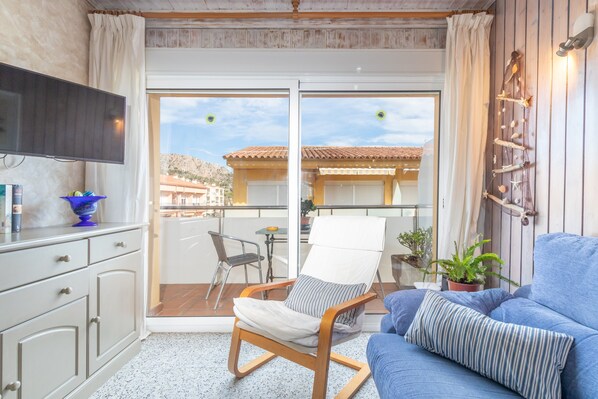 Une pièce comprenant une télévision, un canapé, un fauteuil, une table à manger et un balcon avec vue sur les immeubles.