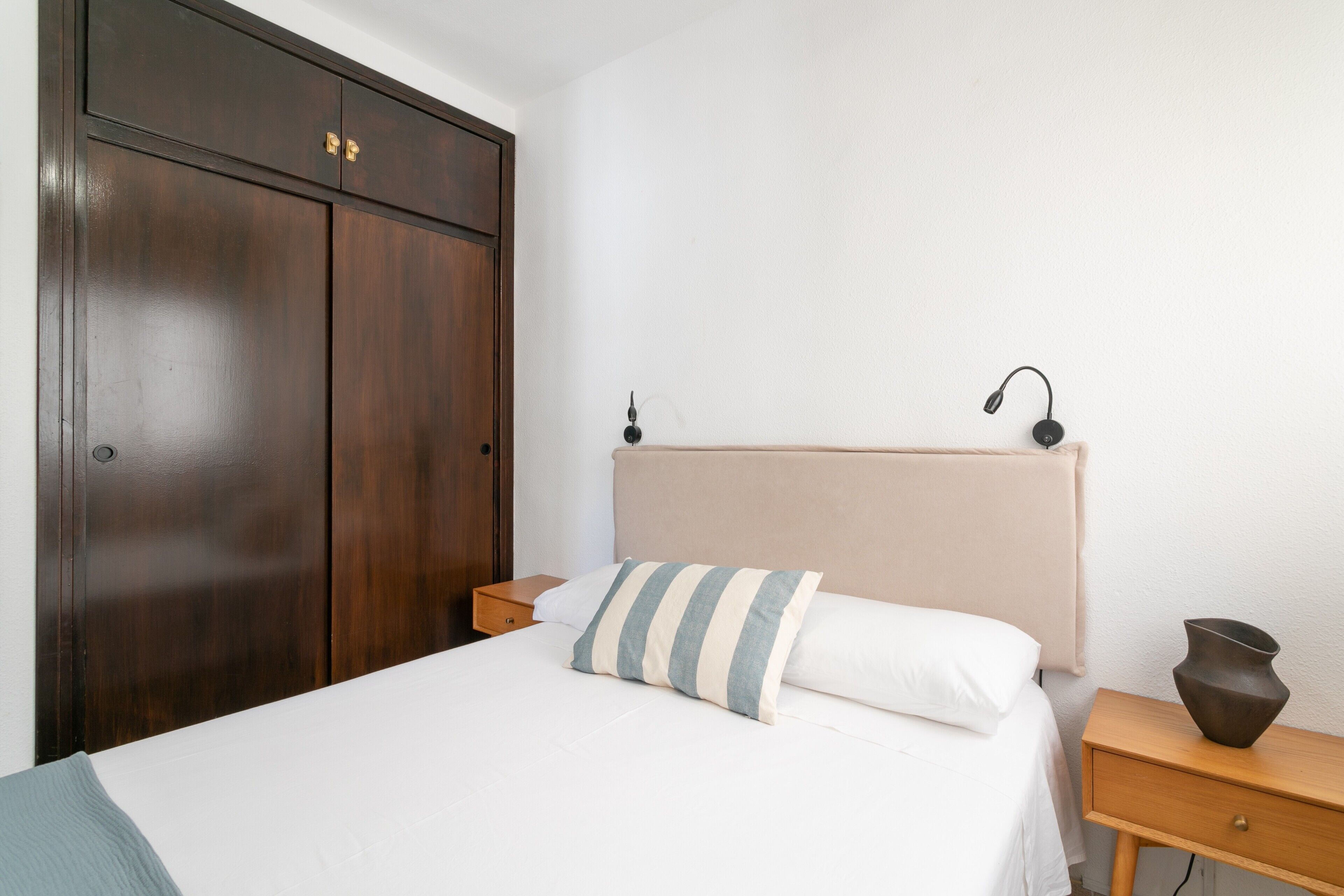Un dormitorio con cama, mesita de noche, armario de madera y lámpara de pared.