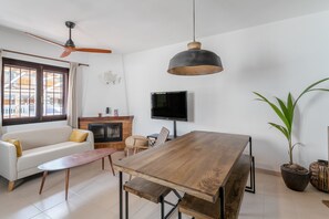 Un salon comprenant un canapé, une table basse, une table à manger en bois, un ventilateur de plafond, un téléviseur, une cheminée et une plante en pot.