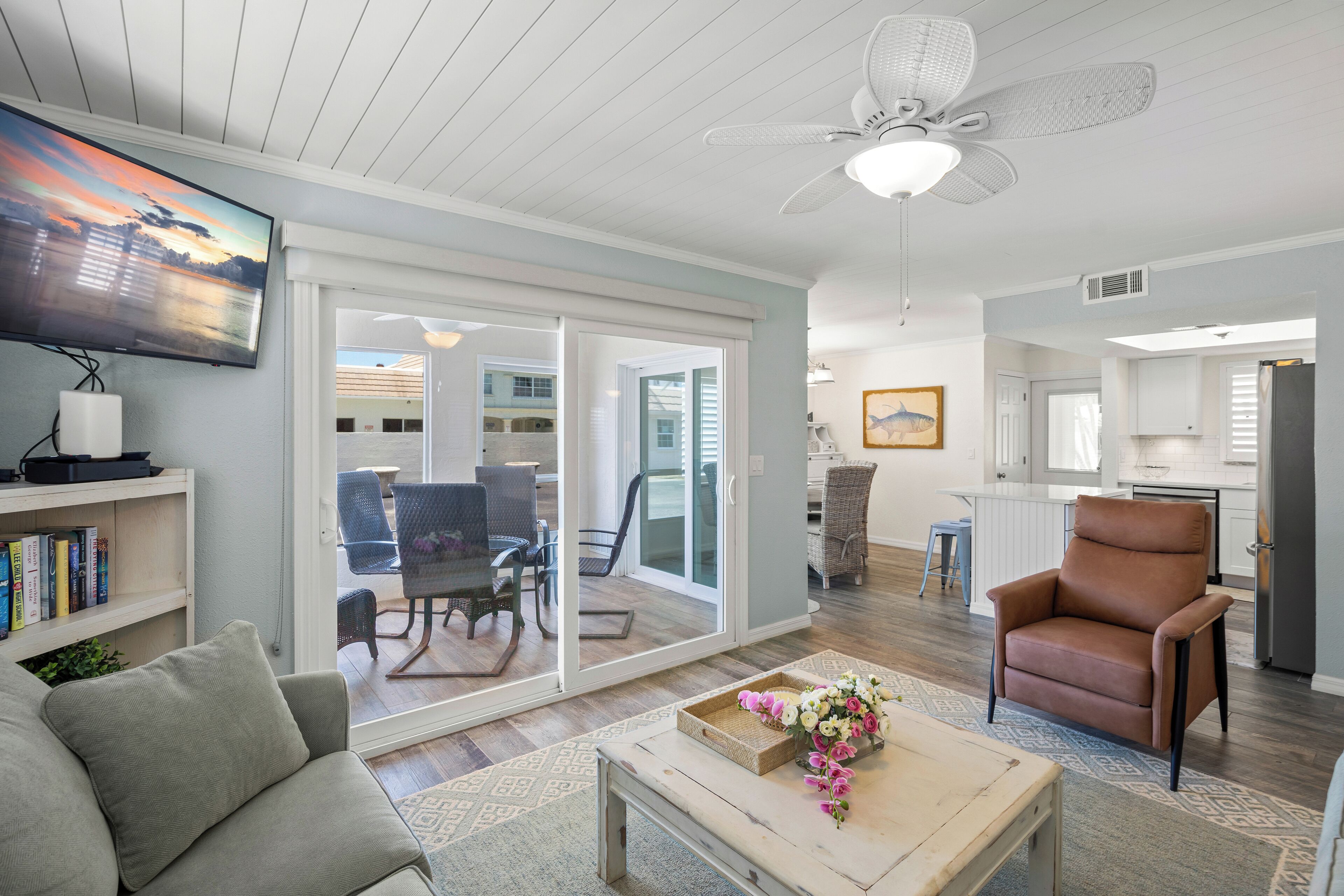 A spacious living room with a flat-screen TV, a comfortable sofa, a coffee table with flowers, and a ceiling fan.
