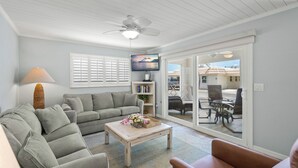 Ein Wohnzimmer mit grauem Sofa, Couchtisch mit Blumen, Fernseher und Deckenventilator.