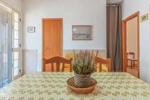 A room with a bed, a dining table with a plant, wooden chairs, a framed picture on the wall, and a door.