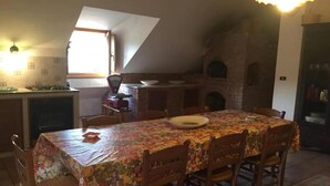 A dining area with a table covered in a floral tablecloth, surrounded by wooden chairs. A brick oven is visible in the background, and a window allows natural light into the room. - Apartment 'Residence Lithos' with Mountain View, Garden and Wi-Fi (Petralia Soprana)