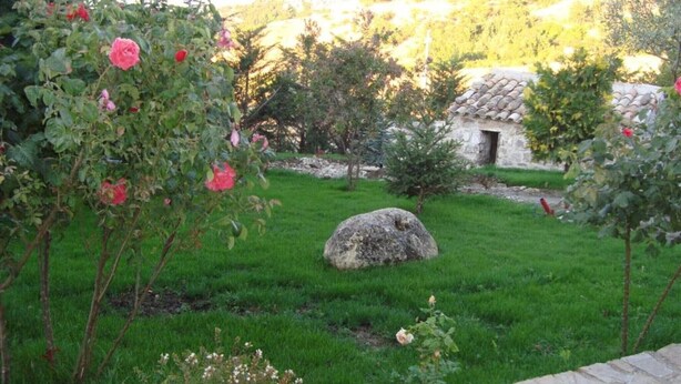 A stone building with a tiled roof, surrounded by a grassy area with a large rock and flowering plants. - Apartment 'Residence Lithos' with Mountain View, Garden and Wi-Fi (Petralia Soprana)