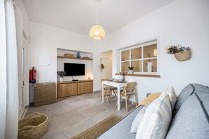 A modern living room with a sofa, a dining table with chairs, a television, and a kitchen area with a counter and cabinets.