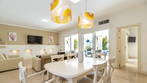 Un espace repas moderne avec une table et des chaises blanches, un téléviseur à écran plat fixé au mur et une vue sur l’extérieur grâce à de grandes fenêtres.