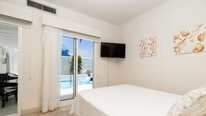 Une chambre avec un lit, un téléviseur fixé au mur et une vue sur une piscine et l’océan, accessible par une porte coulissante.
