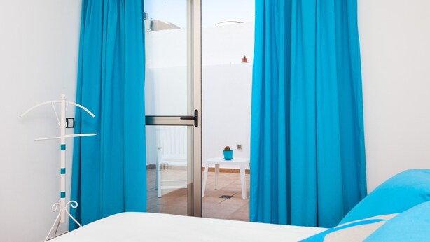 A room with a bed, a white chair, a small table with a potted plant, and blue curtains. - Vacation Home "Corralejo Main Street" with Terrace & Wi-Fi (Corralejo)