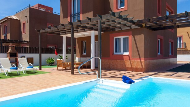 Una piscina con muelle de madera, sillas de descanso y una pérgola.