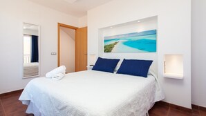 Habitación de hotel con una cama grande, un cuadro de una playa tropical, un espejo y una mesita de noche con lámpara.