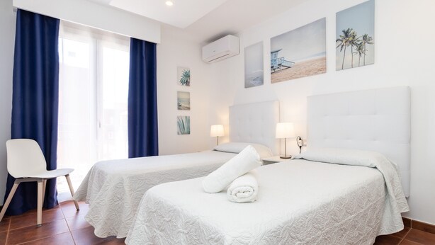 A hotel room with two beds, a chair, a window with curtains, wall art, and an air conditioning unit.