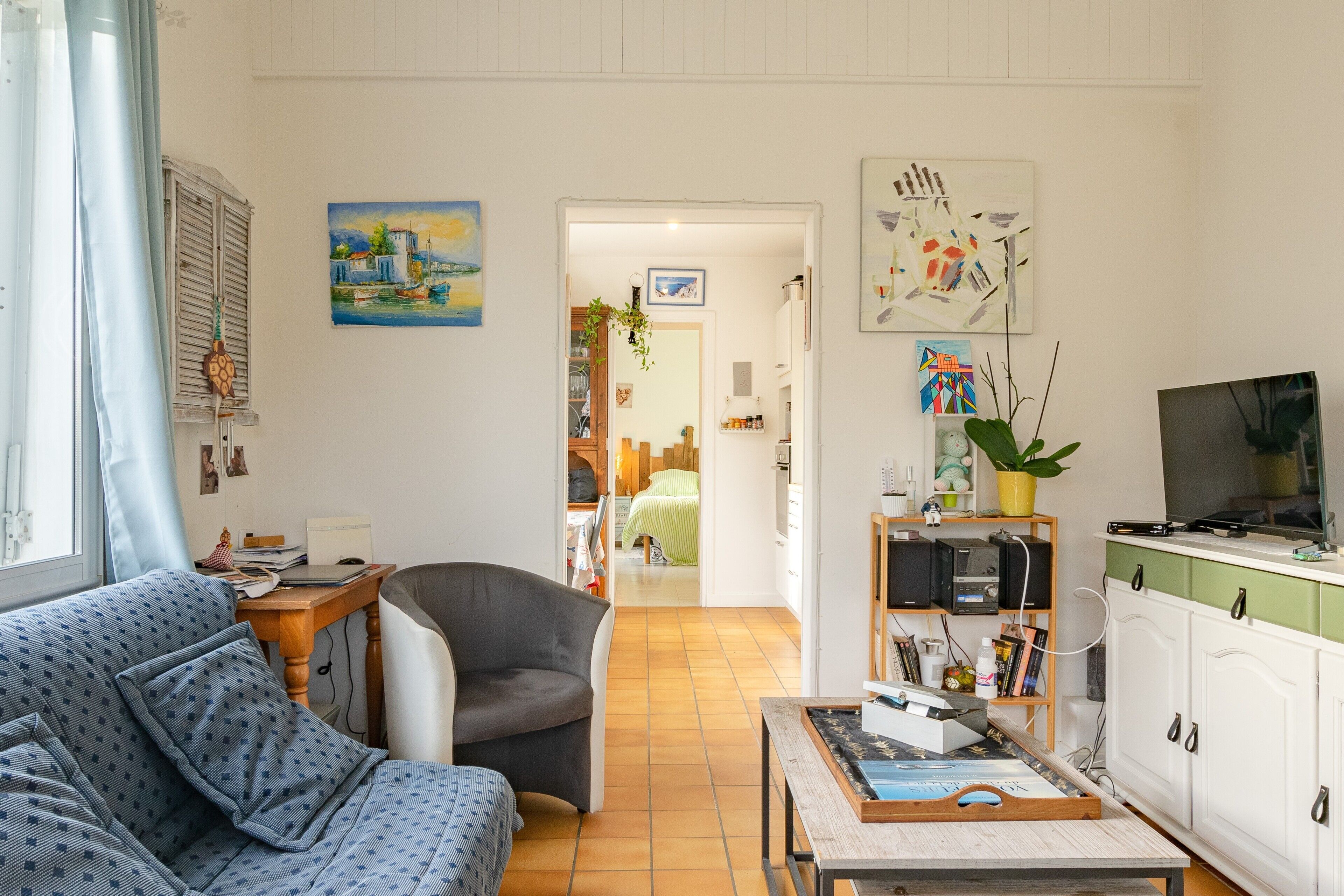 Un soggiorno con divano, poltrona e un tavolo con libri e una pianta. È presente una zona cucina con pensili e un televisore.