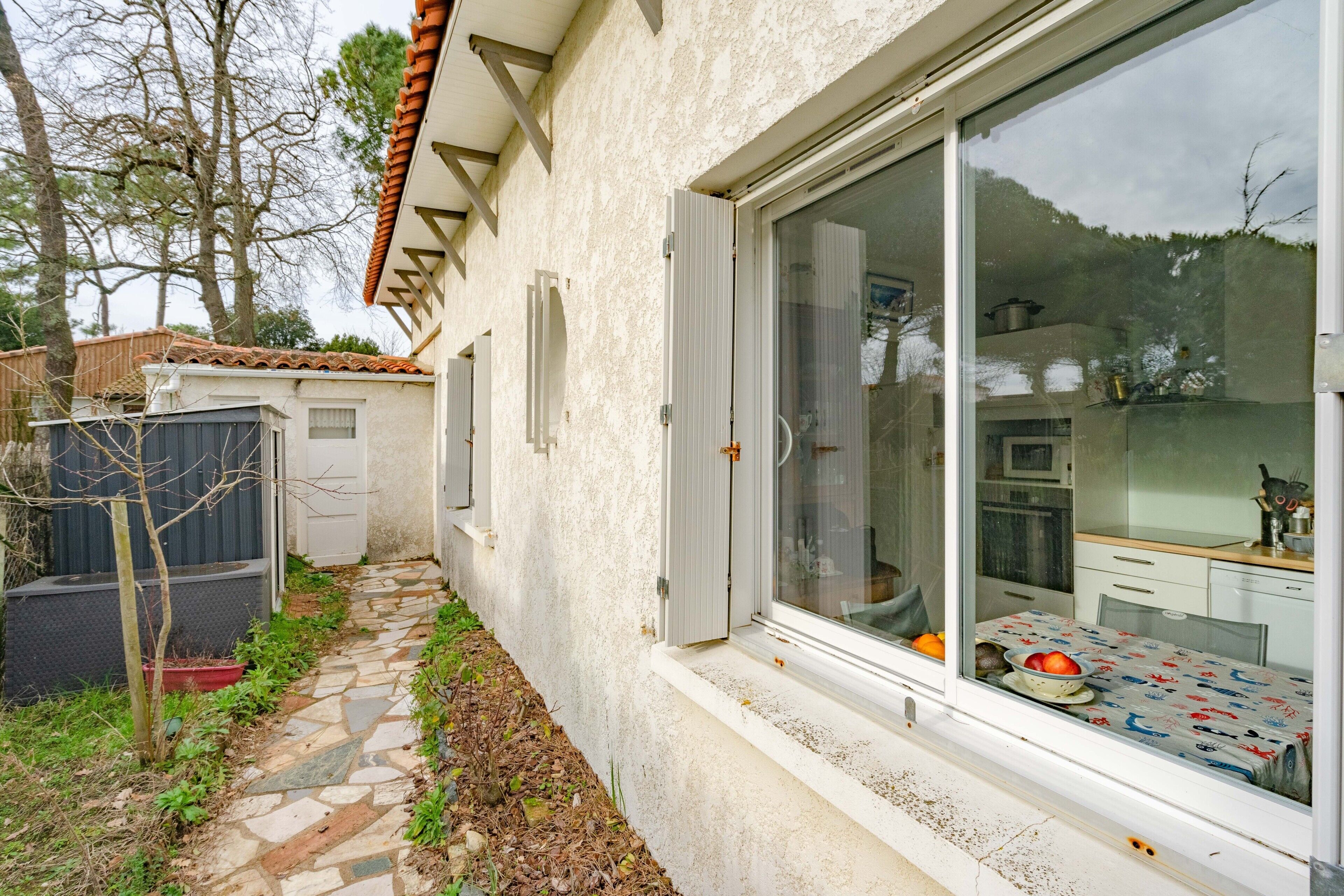 Un bâtiment blanc avec un toit recouvert de tuiles, un chemin de pierre et une fenêtre donnant sur une table et des fruits.