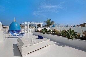 Une terrasse sur le toit avec des chaises longues blanches, un espace salon couvert et des structures surmontées de dômes bleus.