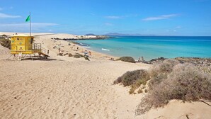 Et redderfyr på en sandstrand med klart, blåt vand og fjerne bjerge.