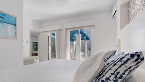 Et soveværelse med en stor seng, hvidt sengetøj, en pude med blå og hvide mønstre samt udsigt til det fri gennem en glasdør.