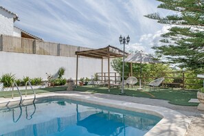 Une piscine agrémentée d’une pergola et de chaises longues, entourée d’un mur blanc et de verdure.