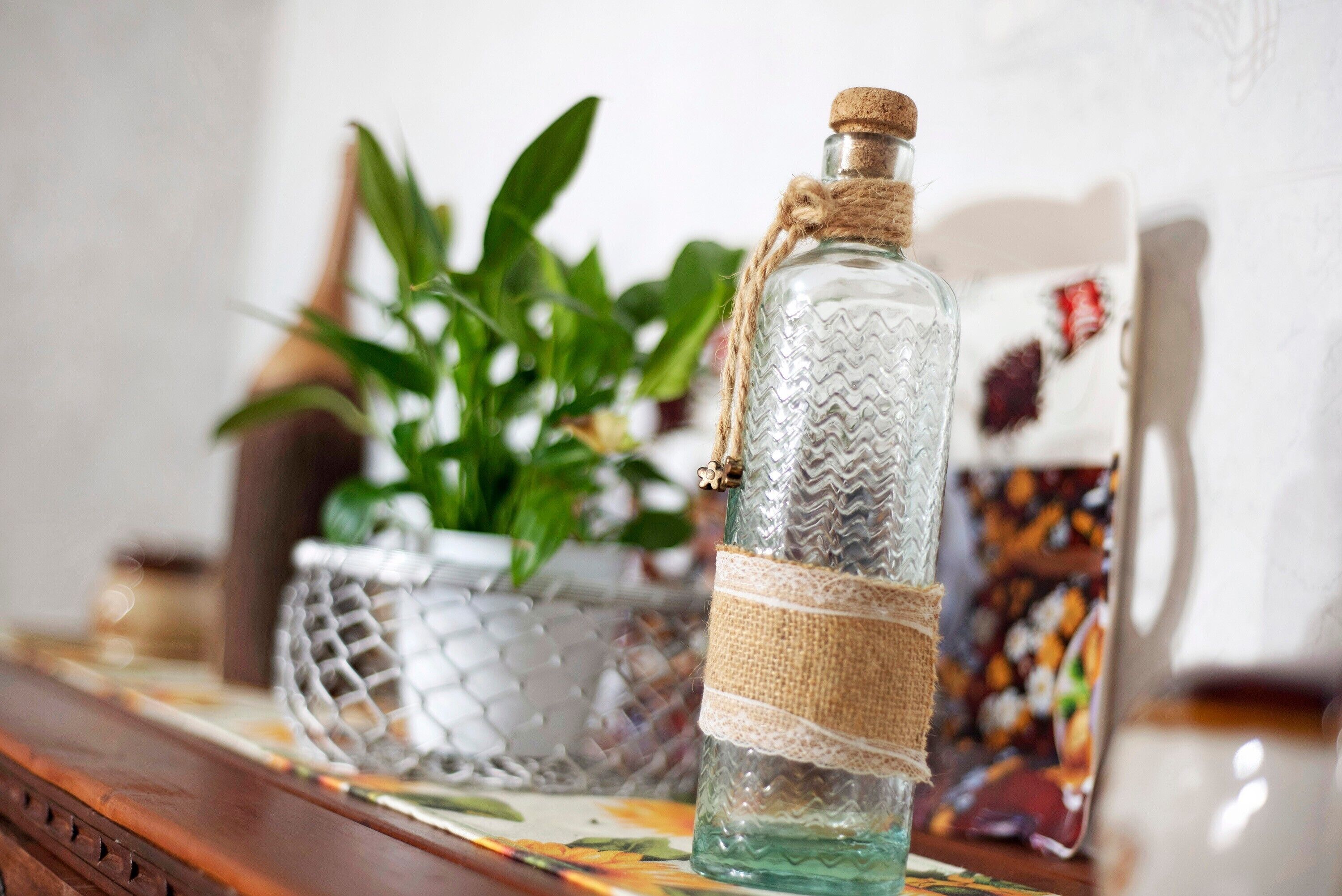 A clear glass bottle with a cork stopper, tied with a burlap ribbon, placed on a table with a woven basket and a potted plant in the background.
