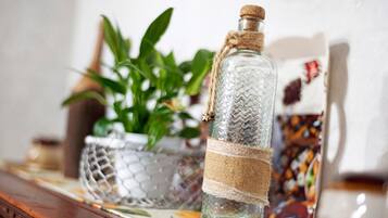 A clear glass bottle with a cork stopper, tied with a burlap ribbon, placed on a table with a woven basket and a potted plant in the background.