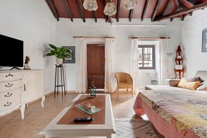 A bedroom with a bed, a TV, a coffee table, a chair, a plant, and a door leading to another room.