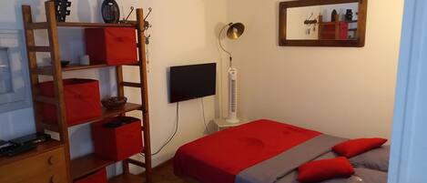 Un dormitorio con cama, televisor, calefactor y estante de madera con cajas rojas.