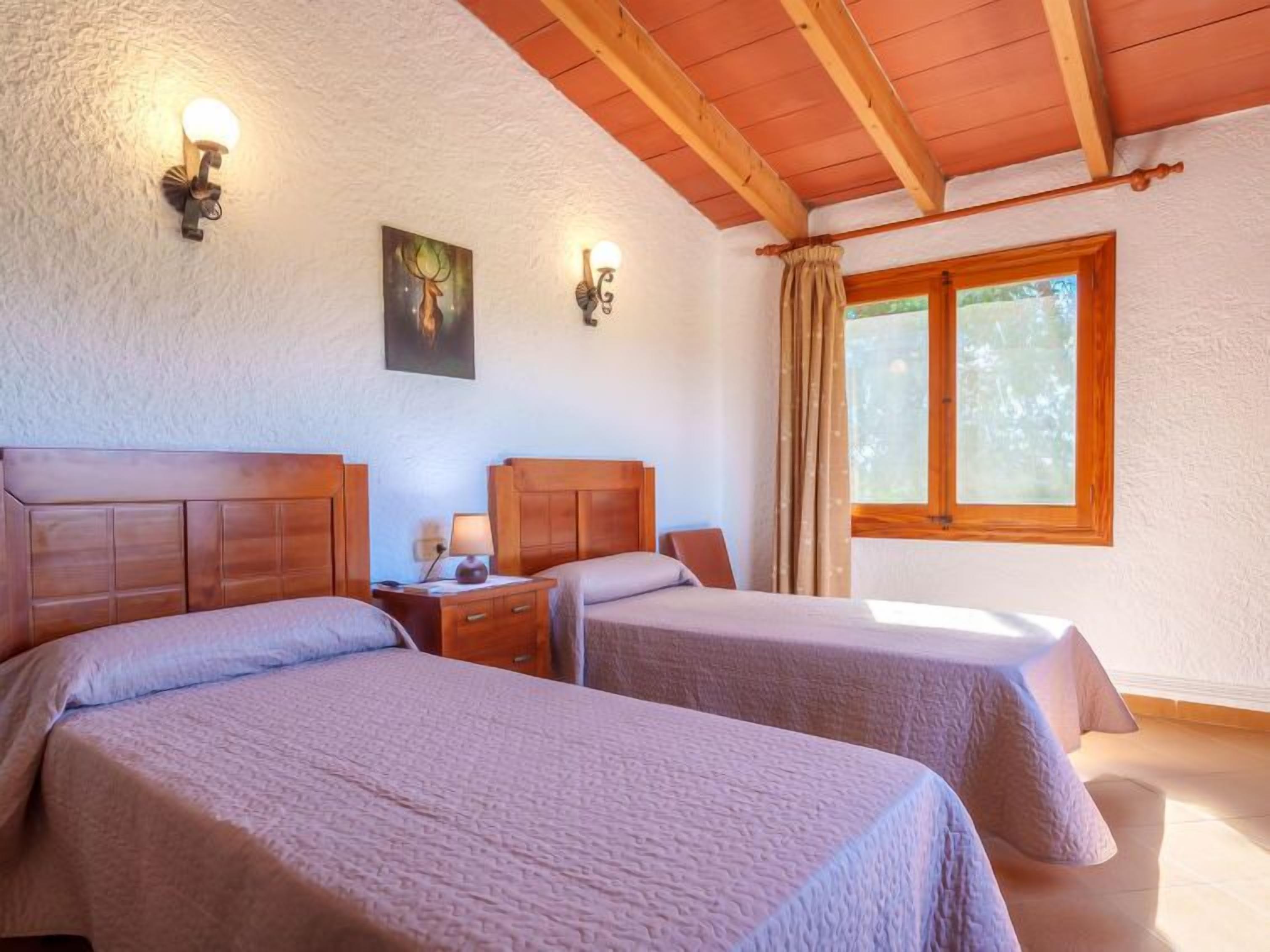 A room with two beds, wooden headboards, a nightstand with a lamp, a painting on the wall, and a window with curtains.