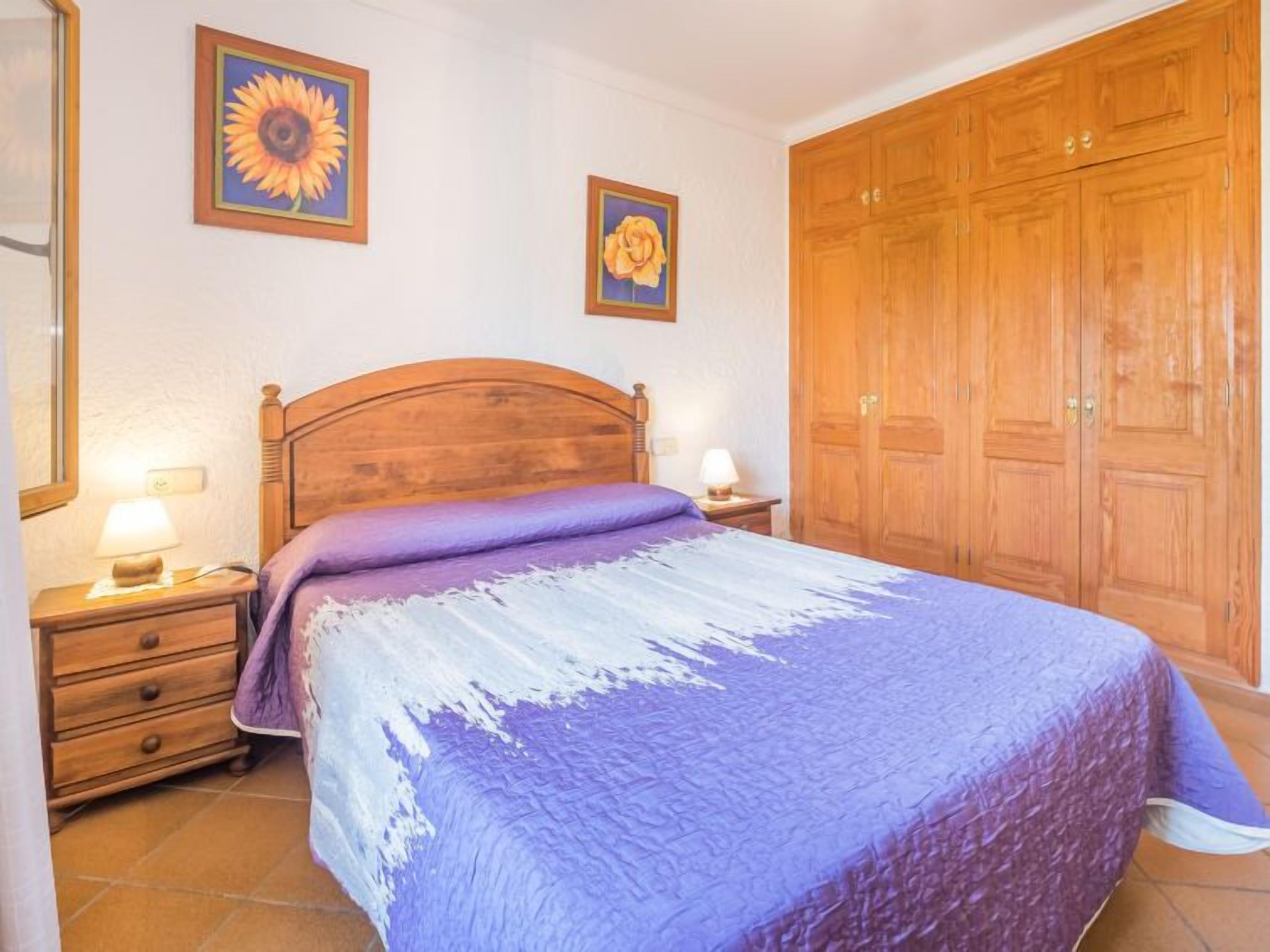 A bedroom with a wooden bed, two bedside tables with lamps, a large wardrobe, and two framed floral paintings on the wall.