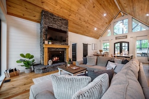 A cozy living room with a stone fireplace, wooden ceiling, and large windows.