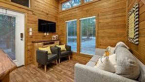 Un intérieur en bois comprenant un canapé, deux fauteuils, un téléviseur et une fenêtre avec des stores.