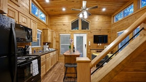 Une cuisine rustique dans une cabane en bois, avec un réfrigérateur, une cuisinière et un four à micro-ondes, tous de couleur noire. On y trouve également un tabouret de bar en bois et un ventilateur de plafond.