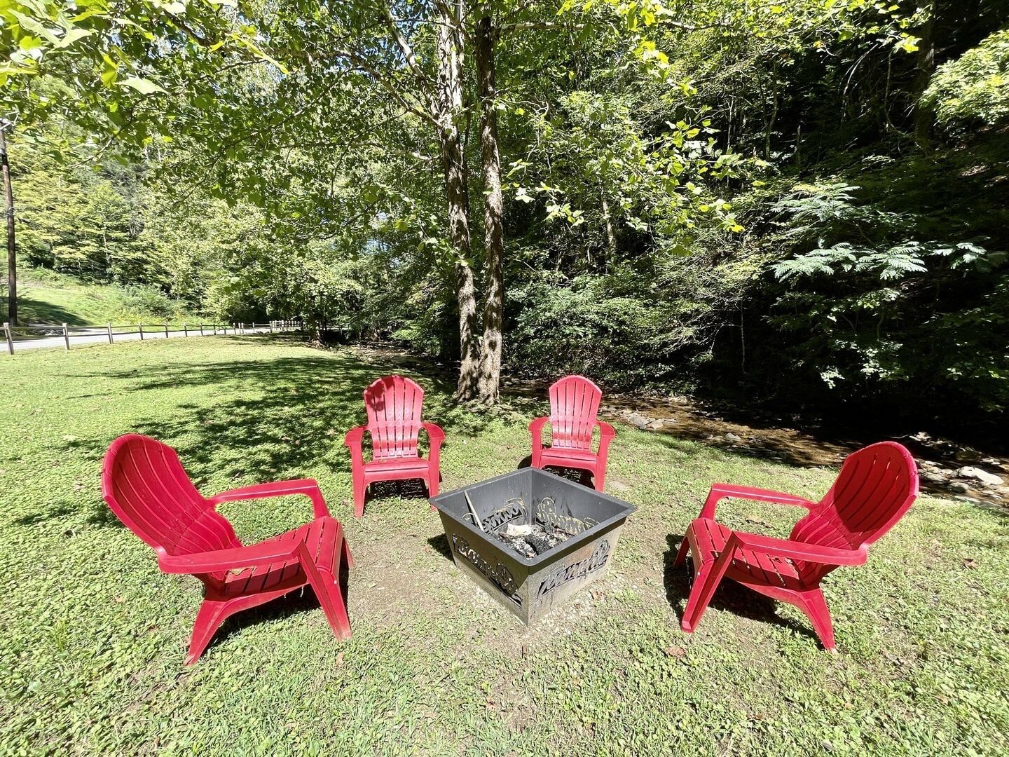 A fire pit surrounded by four red Adirondack chairs in a grassy outdoor area with trees.