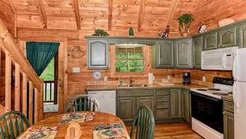 A wooden cabin interior with a kitchen, dining table, chairs, and a window with a view of greenery.