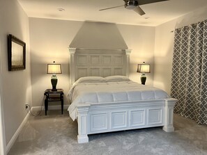 Quarto com cama branca, duas luminárias de cabeceira, ventilador de teto e cortina estampada.