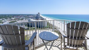 A balcony with two Adirondack chairs and a small table, overlooking a beach and cityscape.