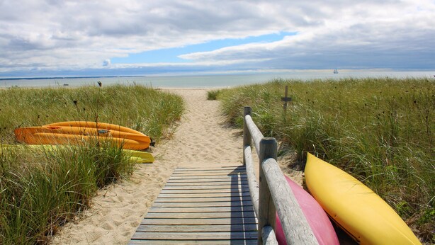 Ein hölzerner Steg führt zu einem Strand, im Hintergrund sind Kajaks und ein Segelboot zu sehen.