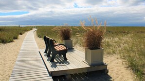 Ein hölzerner Steg führt zu einer Bank und Kübelpflanzen, mit einem Sandstrand und grünen Dünen im Hintergrund.