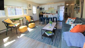 Eine moderne Küche mit weißen Schränken, einem Edelstahlkühlschrank und einer Mikrowelle. Es gibt einen Essbereich mit Tisch und Stühlen sowie ein Wohnzimmer mit einer Couch und Poufs.