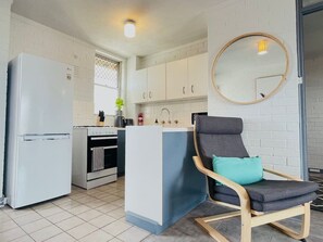 Une cuisine avec des meubles blancs, un réfrigérateur, un four et un évier. On y trouve une chaise avec un coussin et un miroir rond accroché au mur.