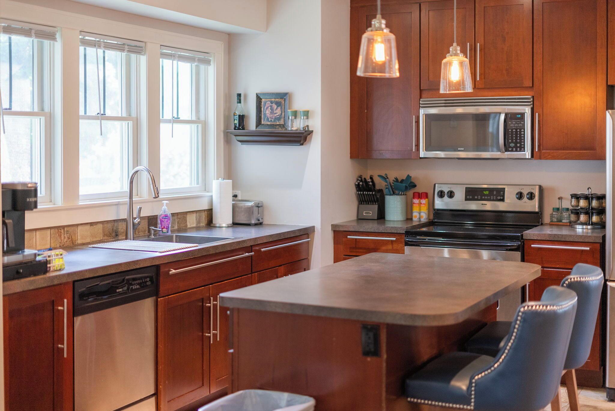 A modern kitchen with wooden cabinets, stainless steel appliances, a built-in island with bar stools, and pendant lighting.