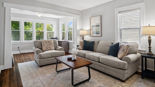 A living room with two sofas, a coffee table, and a lamp. There are large windows with blinds, a ceiling light, and a framed picture on the wall.