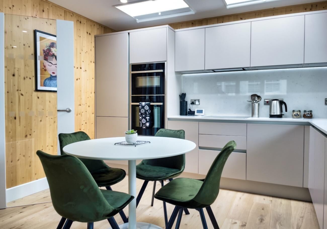 A modern kitchen with white cabinets, a round dining table with green chairs, and a wooden accent wall.