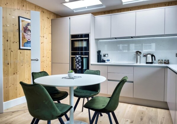 A modern kitchen with white cabinets, a round dining table with green chairs, and a wooden accent wall.
