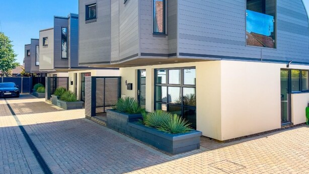 Une maison moderne à deux étages, avec une allée pavée et une voiture bleue garée devant.