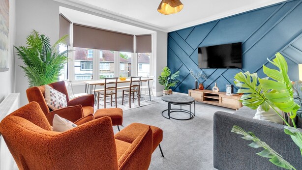 Un salon moderne avec un canapé, des fauteuils, une table basse et un téléviseur fixé sur un mur bleu.
